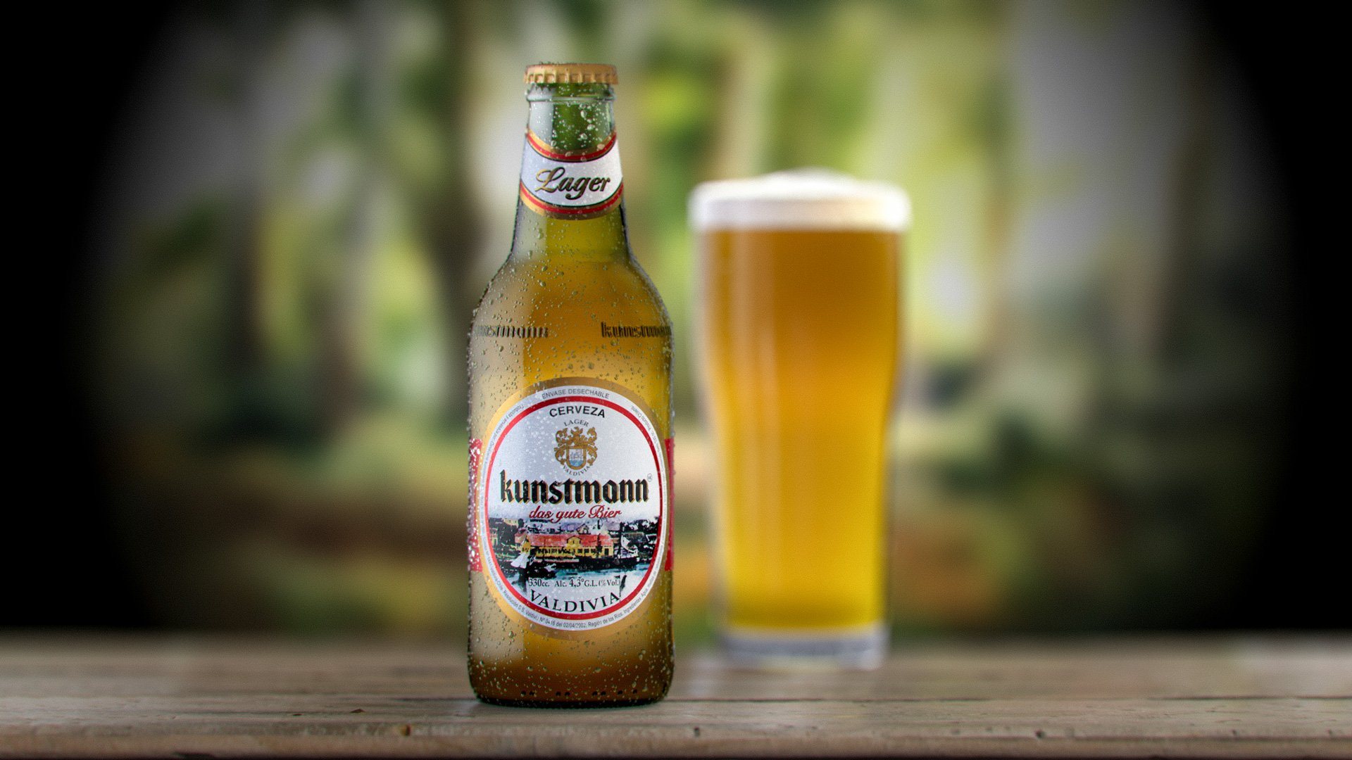 Botella de Kunstmann Lager y un vaso servido, en render 3D sobre un fondo de bosque desenfocado.