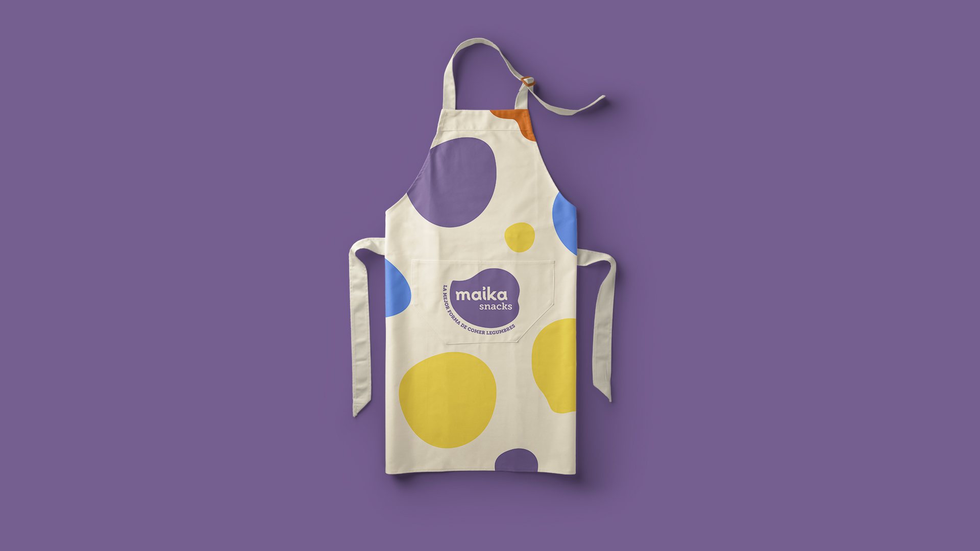 Diseño de delantal crudo de Maika con manchas violeta, amarilla y azul, y el isologo con la frase «La mejor forma de comer legumbres» en el bolsillo.