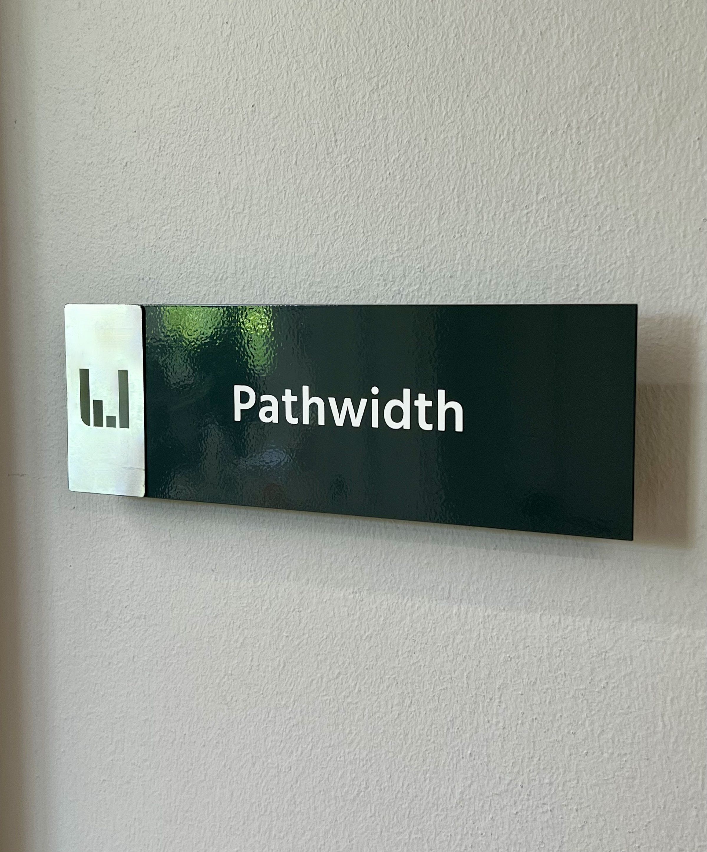 Señalética de sala «Pathwidth» en verde oscuro con el isotipo en metal.
